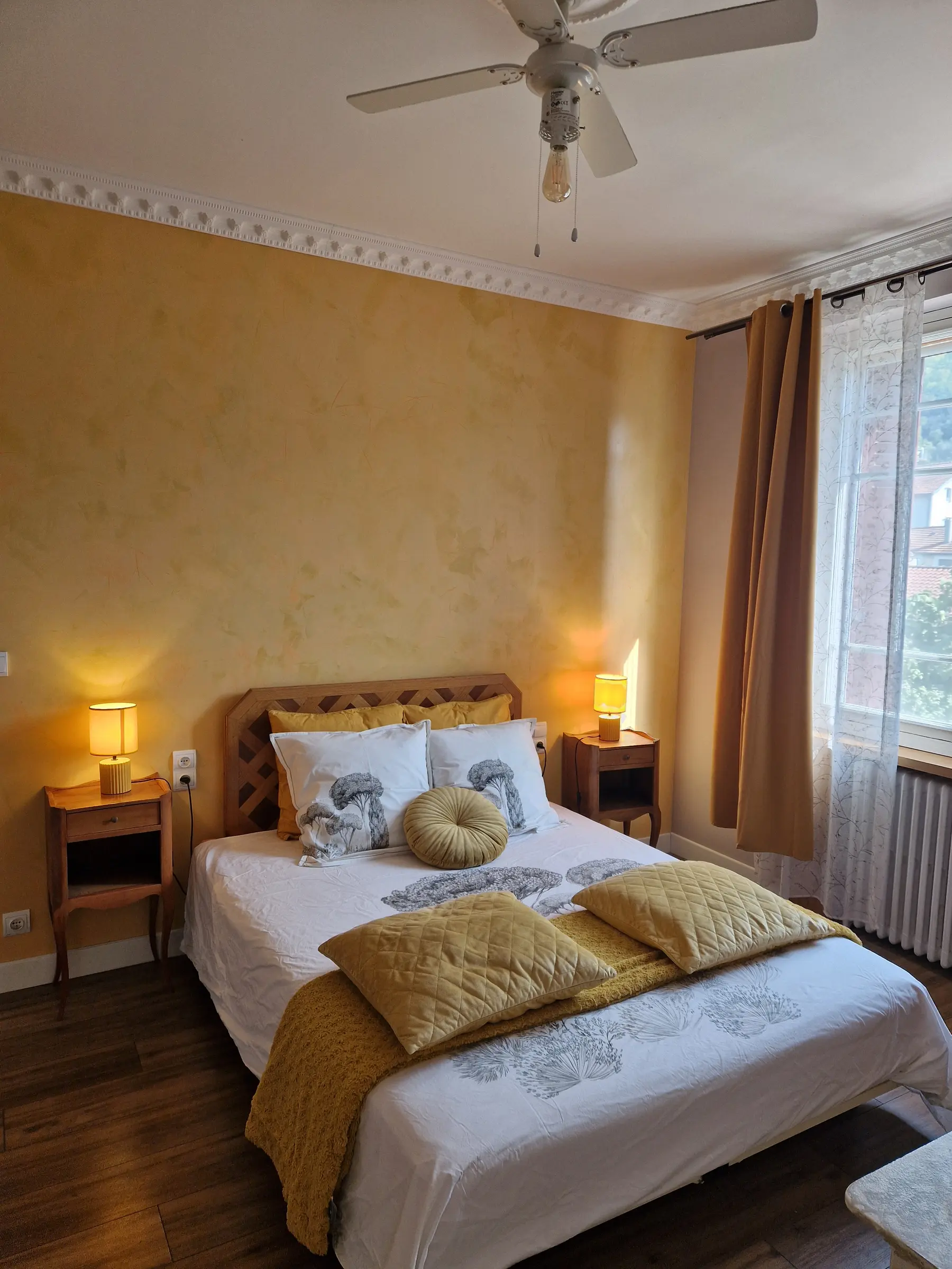 Chambre aux murs jaunes : lit queen, lampes de chevet, ventilateur au plafond et grande fenêtre
