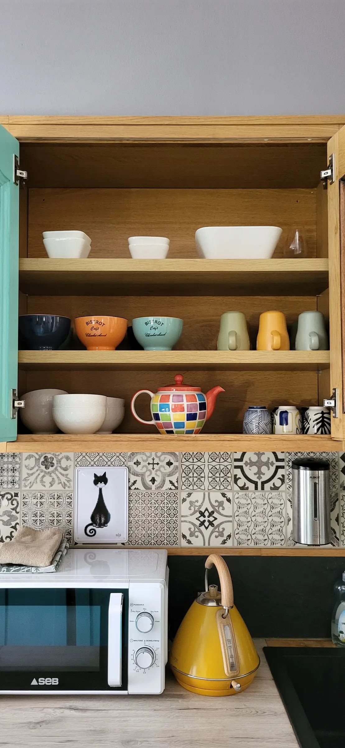 Placard de cuisine ouvert : bols colorés, tasses, théière à carreaux, et bouilloire jaune sur le plan de travail
