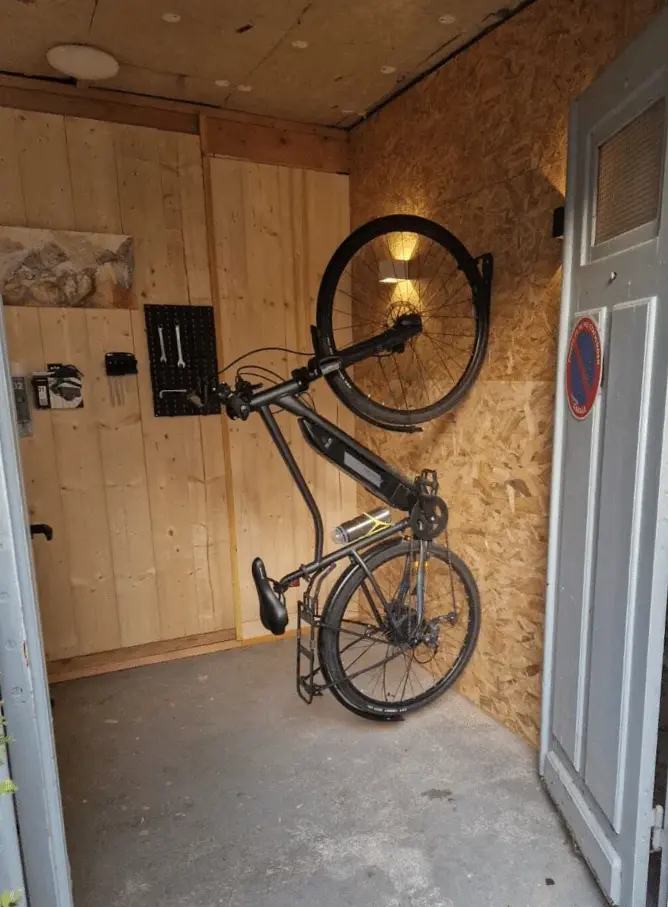 Le local vélo : un vélo suspendu au mur, outils et prises de recharge, murs en bois