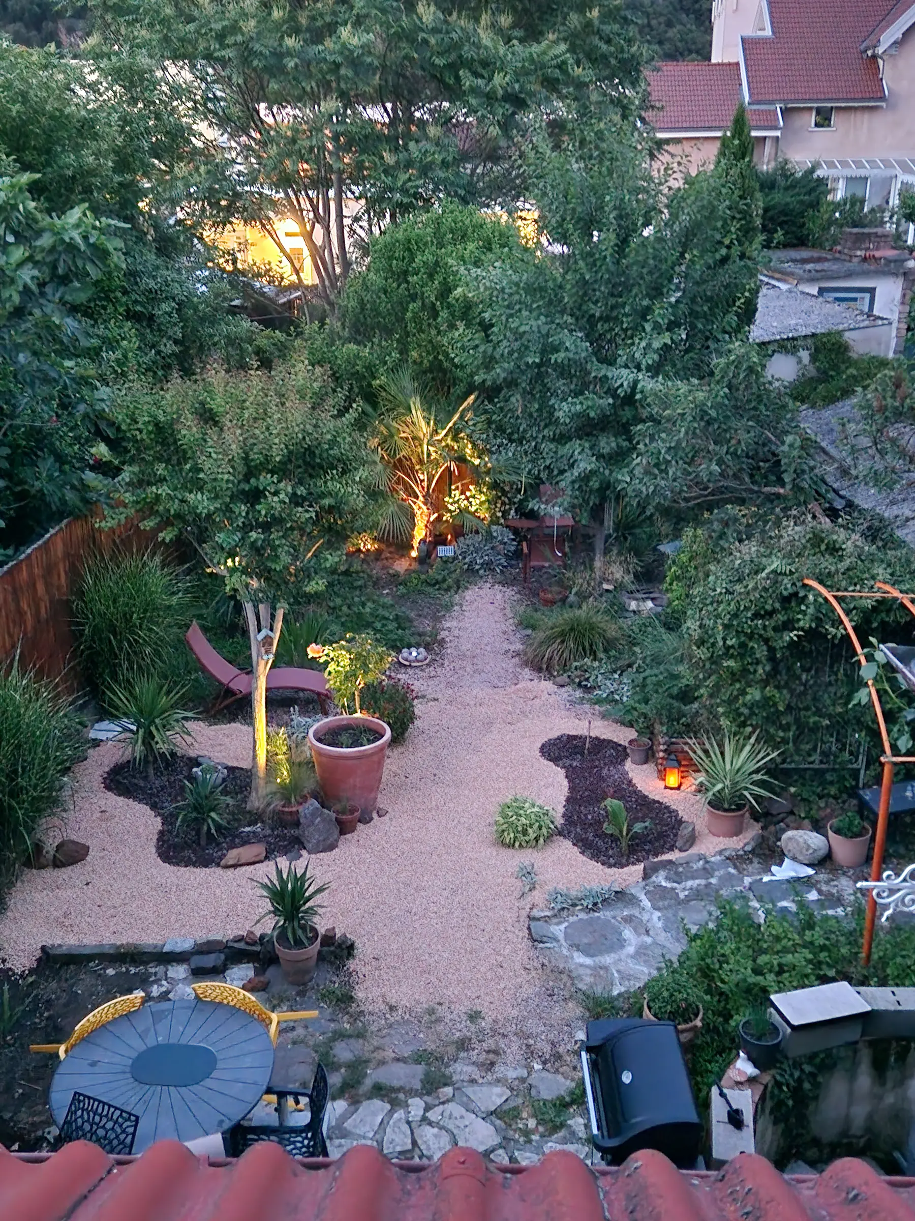 Le jardin vu depuis l'étage au crépuscule : allée de gravier, palmier éclairé, transats et table ronde